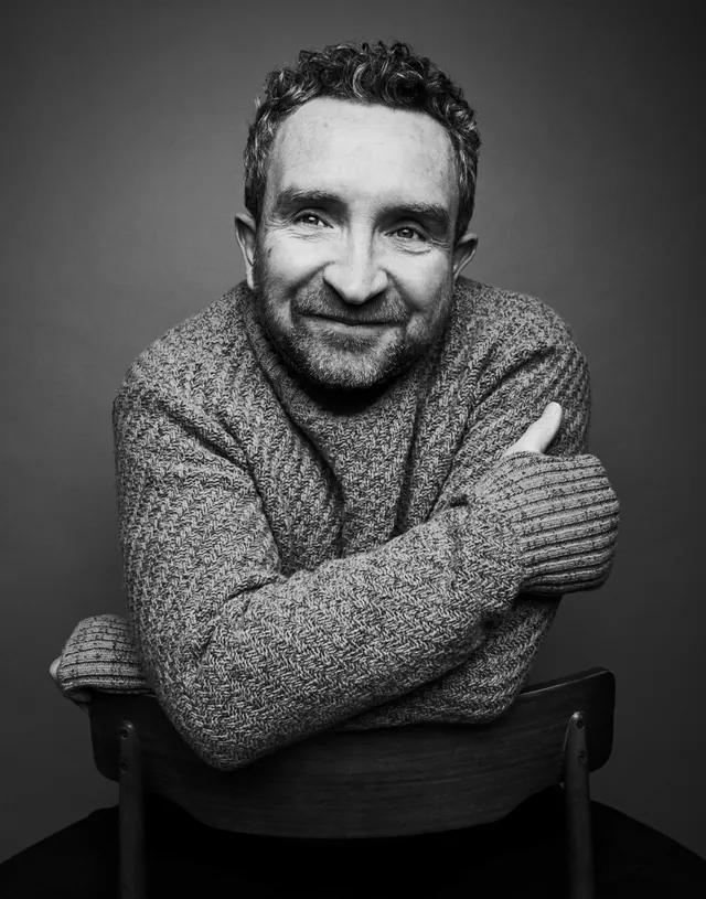 Black-and-white portrait of a smiling actor in a knitted jumper, arms folded over the back of a chair